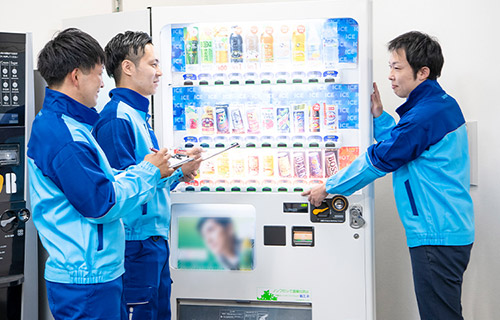 自動販売機の前で先輩社員が教え、後輩社員2名がメモを取る様子